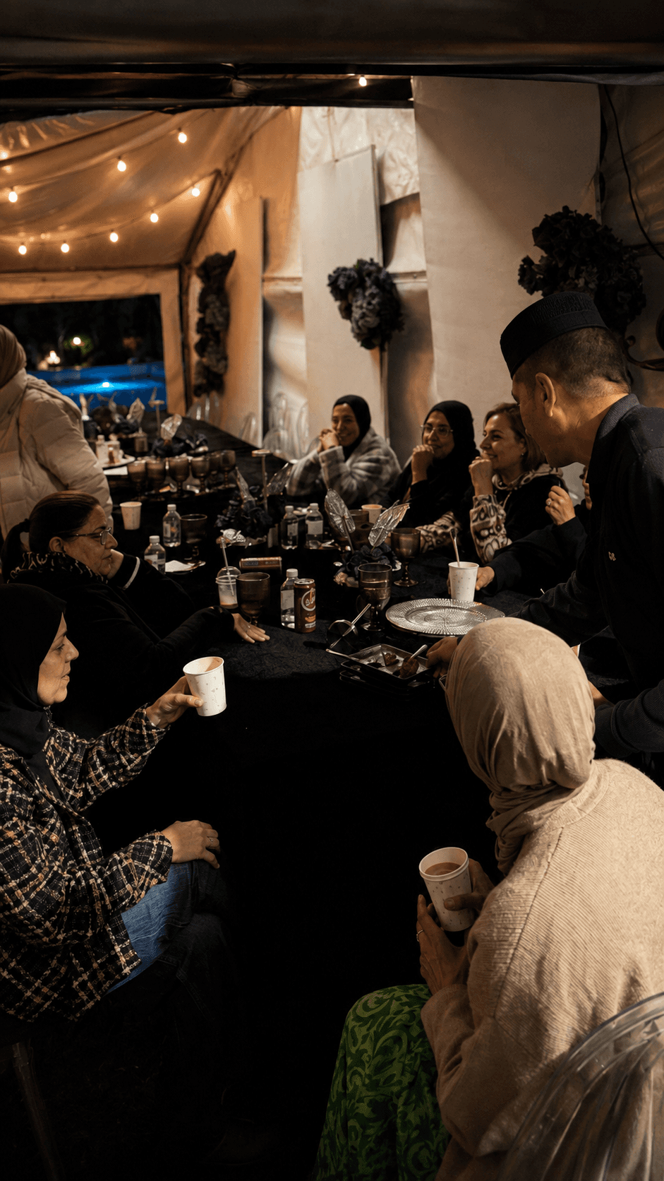 A mixed gathering under the marquee at night — several generations around one table, coffee among them.